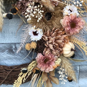 May include: A seasonal wreath with dried flowers, pinecones, and miniature pumpkins in brown, beige, and rose hues. The wreath is set on a natural grapevine base, offering a rustic, autumnal aesthetic. The arrangement includes various textures and tones.