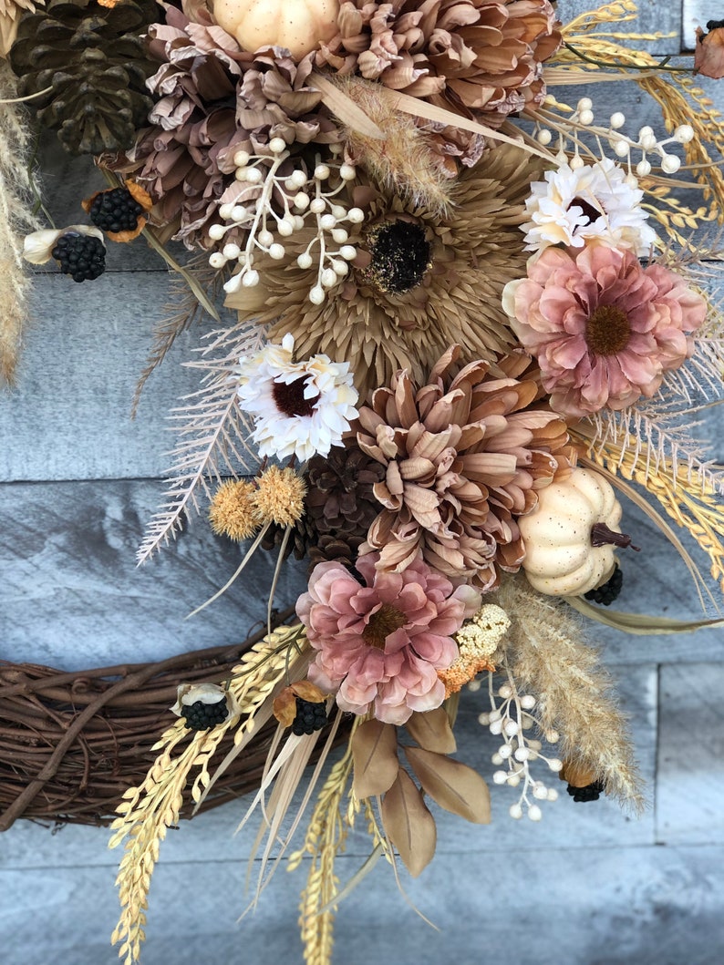 May include: A seasonal wreath with dried flowers, pinecones, and miniature pumpkins in brown, beige, and rose hues. The wreath is set on a natural grapevine base, offering a rustic, autumnal aesthetic. The arrangement includes various textures and tones.