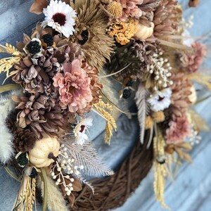 May include: A seasonal wreath with a brown, woven base, decorated with dried flowers, wheat, and miniature pumpkins. The wreath incorporates a palette of neutral tones, such as beige, brown, and hints of white and peach, offering a rustic, autumnal appearance.