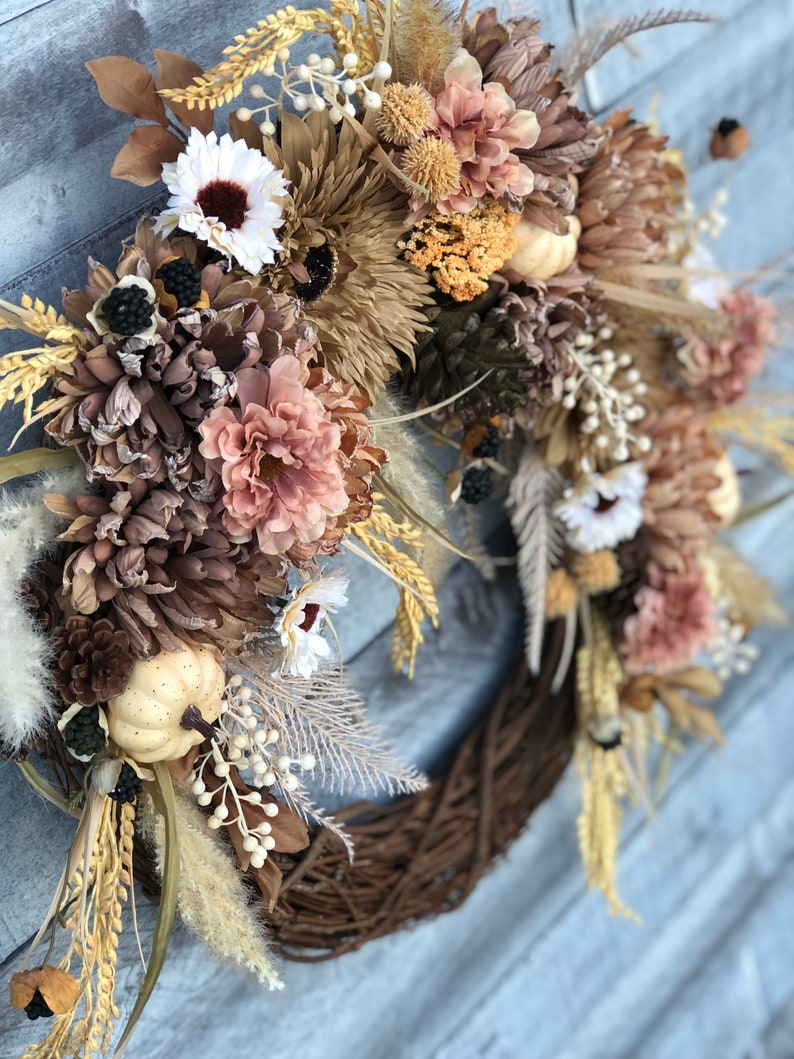 May include: A seasonal wreath with a brown, woven base, decorated with dried flowers, wheat, and miniature pumpkins. The wreath incorporates a palette of neutral tones, such as beige, brown, and hints of white and peach, offering a rustic, autumnal appearance.