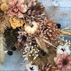 May include: A seasonal wreath with dried flowers, wheat, and pinecones in brown, beige, and cream hues. A small, cream-coloured pumpkin is placed amongst the flowers. Ideal for autumn home decoration, the wreath has a rustic aesthetic.