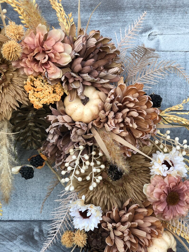 May include: A seasonal wreath with dried flowers, wheat, and pinecones in brown, beige, and cream hues. A small, cream-coloured pumpkin is placed amongst the flowers. Ideal for autumn home decoration, the wreath has a rustic aesthetic.