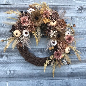 May include: A seasonal wreath with a brown, woven base. It features artificial flowers in brown, cream, and tan hues, alongside miniature white pumpkins, pine cones, and wheat. The backdrop is a weathered, grey wooden wall.