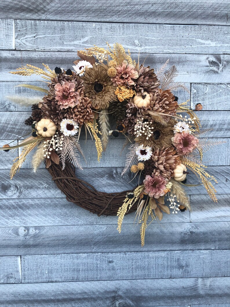 May include: A seasonal wreath with a brown, woven base. It features artificial flowers in brown, cream, and tan hues, alongside miniature white pumpkins, pine cones, and wheat. The backdrop is a weathered, grey wooden wall.