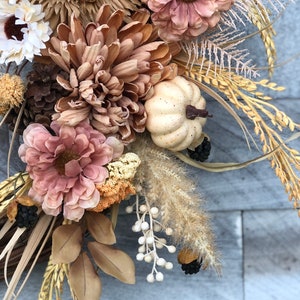 May include: A seasonal wreath with faux flowers in brown, beige, and pink. It features a small white pumpkin, pine cones, and wheat stalks. The design evokes a rustic, autumnal feel, perfect for home decor.