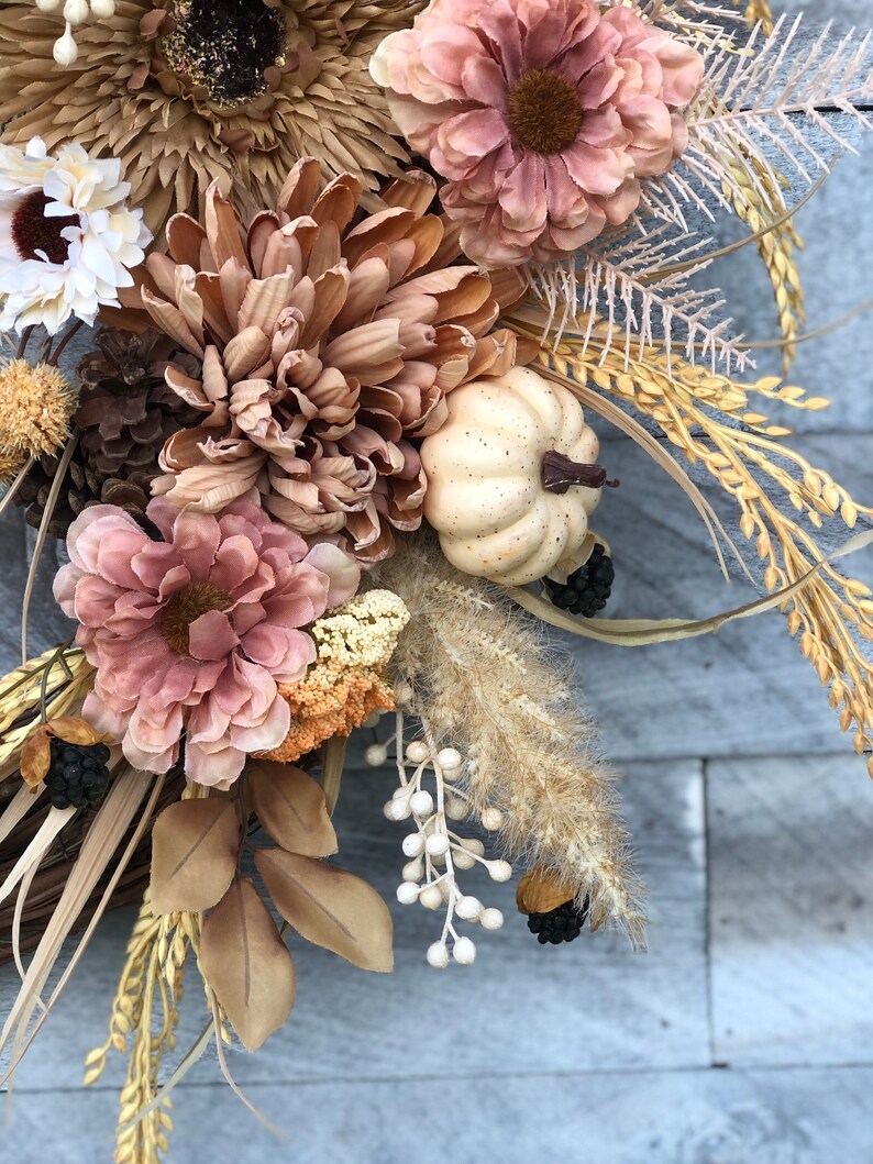 May include: A seasonal wreath with faux flowers in brown, beige, and pink. It features a small white pumpkin, pine cones, and wheat stalks. The design evokes a rustic, autumnal feel, perfect for home decor.