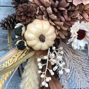 May include: A seasonal wreath with a cream pumpkin, pine cones, and dried flowers in brown, beige, and cream hues. The wreath is set on a woven base, offering a rustic, natural look. The arrangement includes various textures and shapes.