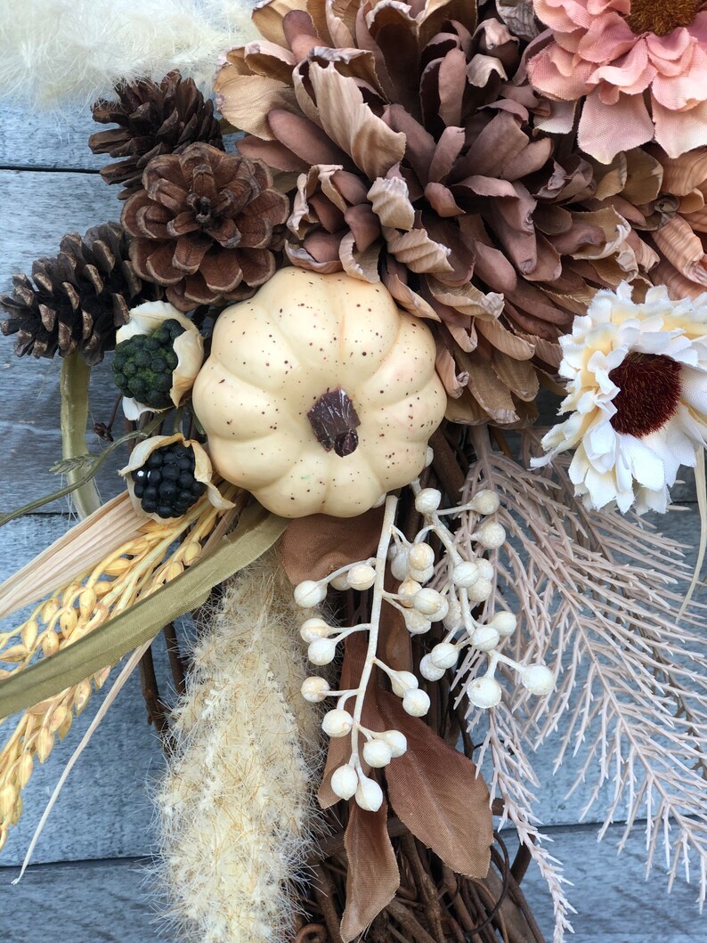 May include: A seasonal wreath with a cream pumpkin, pine cones, and dried flowers in brown, beige, and cream hues. The wreath is set on a woven base, offering a rustic, natural look. The arrangement includes various textures and shapes.