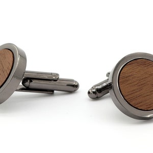 Peut inclure: Une paire de boutons de manchette avec une base ronde en métal gris foncé et un incrustation en bois. L'incrustation en bois est de couleur brune et présente un motif de grain de bois.