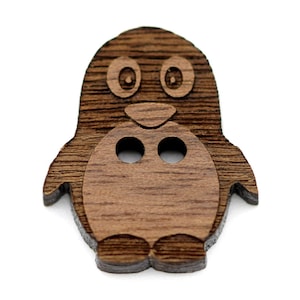 Puede incluir: Un botón de madera con forma de pingüino con dos agujeros para coser. El botón es marrón y tiene un diseño detallado.