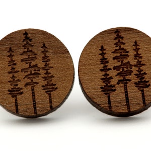 Puede incluir: Un par de pendientes de madera con un diseño de tres pinos grabado con láser. Los pendientes son de color marrón y tienen forma circular.