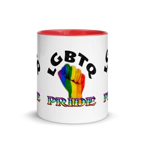 Puede incluir: Taza de cerámica blanca con interior rojo. La taza presenta la palabra "LGBTQ" en negro, un puño de colores del arcoíris y la palabra "PRIDE" en colores del arcoíris. La taza es un símbolo del orgullo LGBTQ+.
