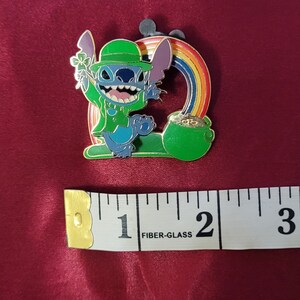 May include: A colorful enamel pin featuring the character Stitch dressed in a green leprechaun outfit, holding a shamrock. A rainbow arches over a pot of gold. The pin measures approximately 2 inches in length.