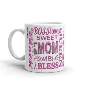 Könnte beinhalten: Weißer Keramikbecher mit rosa und schwarzem Design. Der Becher zeigt das Wort „MOM“ in großen rosa Buchstaben, umgeben von Wörtern wie „BOSS“, „SWEET“ und „HUMBLE“. Ein rosa Band und der Text „800-USH“ sind ebenfalls sichtbar.