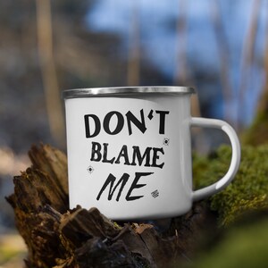 May include: White enamel mug with a silver rim and handle, featuring the text "DON'T BLAME ME" in bold black letters. The mug is set against a blurred natural background, suggesting an outdoor setting. The mug has a simple, classic design.