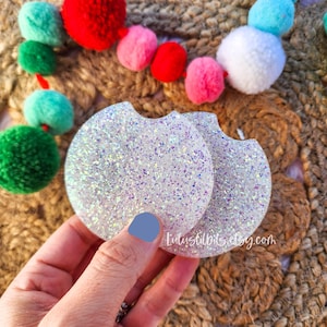 May include: Two white iridescent glitter round drink coasters. The coasters are made of resin and have a smooth, glossy finish.