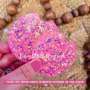 May include: Two pink glitter car coasters with a unique shape. The coasters have a sparkly, textured appearance with purple and yellow accents. The text "luluslitbits.etsy.com" is visible on the coasters. The bottom of the image has the text "*COLORS MAY APPEAR DARKER OR BRIGHTER DEPENDING ON YOUR SCREEN*"