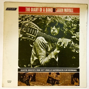 May include: A vintage record album cover featuring a black and white photo of John Mayall holding a guitar. The album title is "The Diary of a Band" and the text "Selected Highspots from Sixty Hours of Bluesbreakers Club Recordings" is printed below the photo. The album cover is white with red and brown accents.