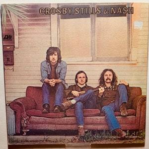 May include: A vintage album cover featuring Crosby, Stills & Nash. The three musicians are sitting on a brown couch in front of a white house with a window. The album cover is a light brown color with a textured surface.