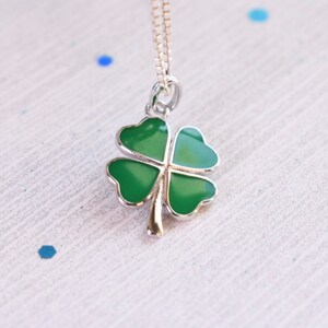 May include: A silver necklace with a four-leaf clover pendant. The clover is finished in bright green enamel with a silver border and a gold-coloured stem. The chain is delicate and hangs from the pendant.