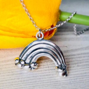 May include: A silver rainbow charm necklace with a chain. The rainbow charm has a textured surface and is detailed with small clouds on each end.