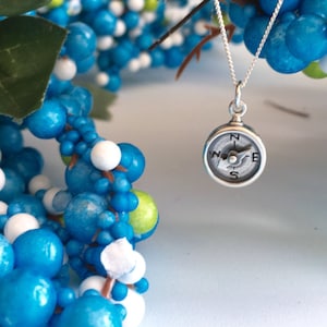 May include: A silver necklace with a compass pendant. The compass is round and has a black face with white markings. The necklace is hanging from a blue and white garland.