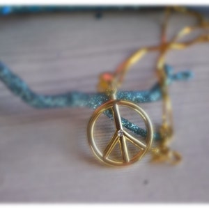 May include: A gold peace sign necklace with a small diamond in the centre. The necklace is on a wooden surface.
