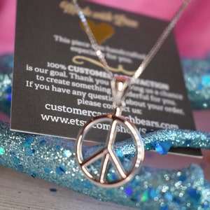 May include: A silver peace sign pendant necklace on a silver chain. The necklace is displayed on a blue glitter surface with a black card behind it. The card has text that says "100% CUSTOMER SATISFACTION is our goal. Thank you for giving us a chance to create something wonderful for you. If you have any questions please contact us: customercachbears.com www.etsy.com"