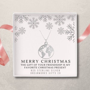 Puede incluir: Un collar de plata con un colgante de mapa del mundo en una caja blanca con un patrón de copo de nieve. La caja tiene el texto "Feliz Navidad, El regalo de tu amistad es mi regalo de Navidad favorito, Plata 925, Dreamworks Gifts Co."