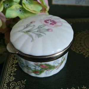 May include: A white porcelain trinket box with a floral design. The lid features a pink carnation with green leaves. The base of the box has a floral pattern with blue and pink flowers.