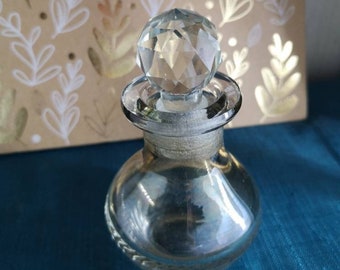 May include: A clear glass bottle with a faceted crystal stopper. The bottle has a round body and a short neck. The stopper is a geometric shape with many facets.