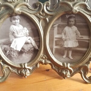 May include: A vintage brass double photo frame with ornate details. Two oval-shaped openings hold black and white photos of young girls.