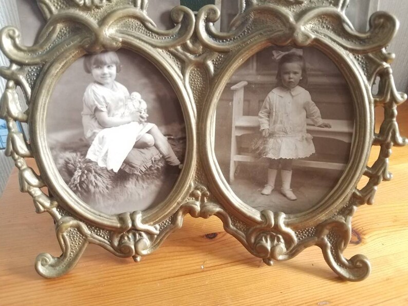 May include: A vintage brass double photo frame with ornate details. Two oval-shaped openings hold black and white photos of young girls.