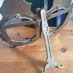 May include: A silver-toned metal picture frame with a decorative design. The frame has two openings and a stand on the back. The stand is adjustable to allow the frame to stand upright.
