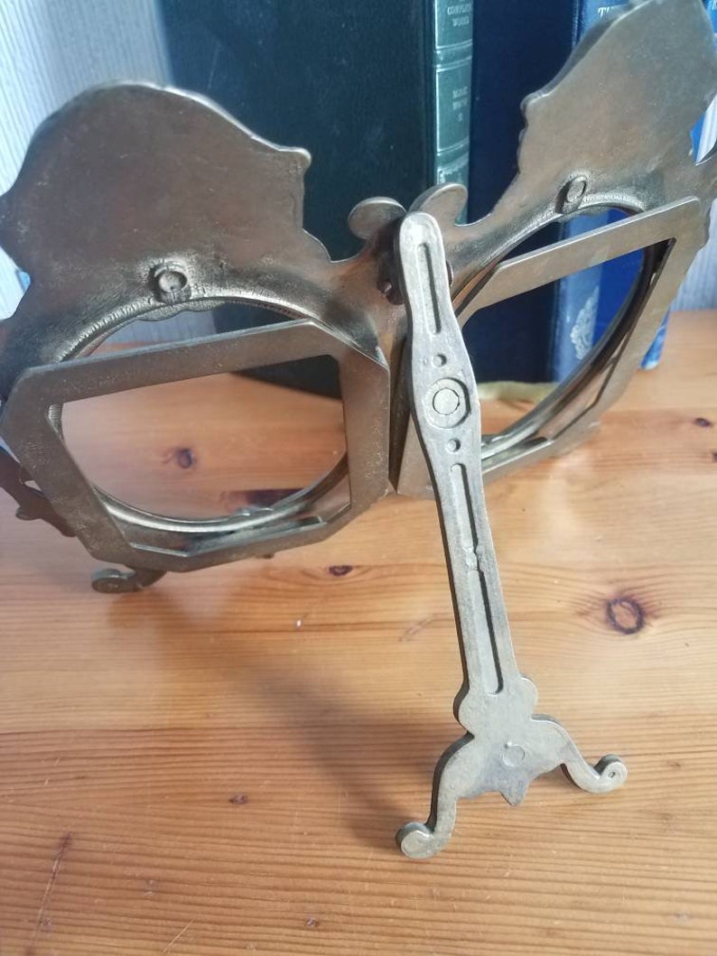 May include: A silver-toned metal picture frame with a decorative design. The frame has two openings and a stand on the back. The stand is adjustable to allow the frame to stand upright.