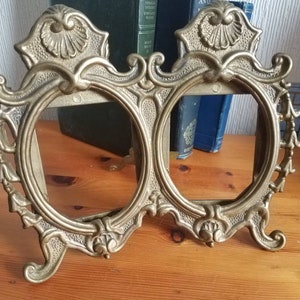 May include: A gold-toned metal double picture frame with ornate details and a scalloped design. The frame has two oval openings and stands on a wooden surface.