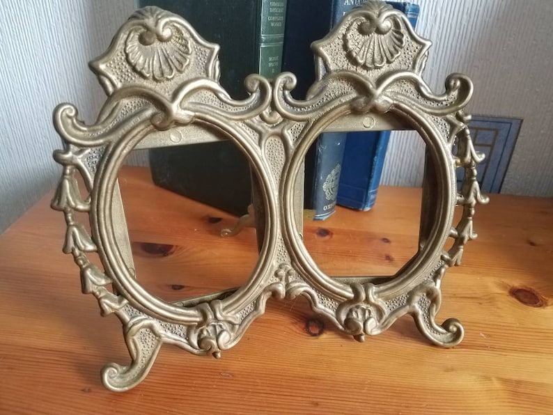 May include: A gold-toned metal double picture frame with ornate details and a scalloped design. The frame has two oval openings and stands on a wooden surface.