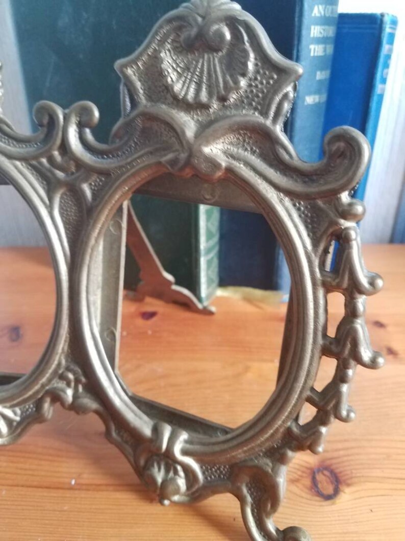 May include: A vintage brass picture frame with ornate details and a decorative design. The frame has two oval openings and a stand on the back.
