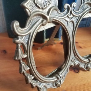 May include: A silver-toned metal picture frame with ornate details and three oval openings. The frame has a decorative top with a shell design and tassels on the sides.