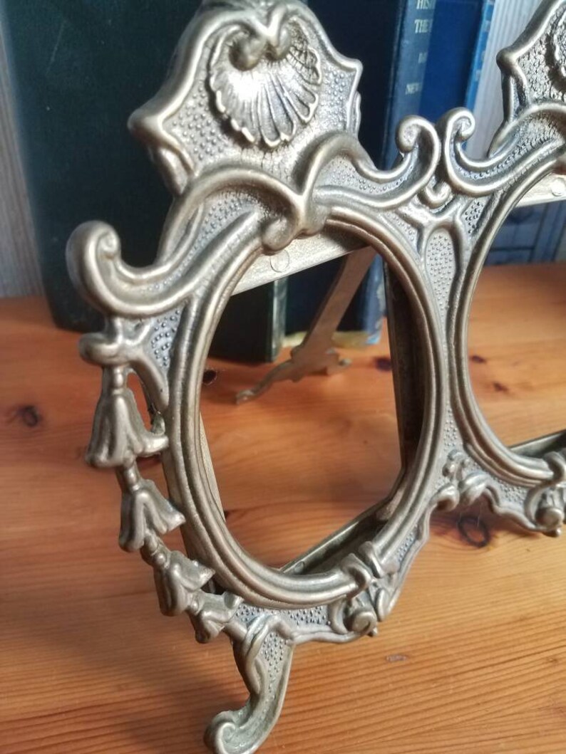 May include: A silver-toned metal picture frame with ornate details and three oval openings. The frame has a decorative top with a shell design and tassels on the sides.