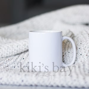May include: A white ceramic mug with a curved handle sits on a textured, off-white knitted blanket. The mug is plain and cylindrical, with a smooth, glossy finish. The blanket has a speckled pattern. The words "kiki's bay design co." are at the bottom.