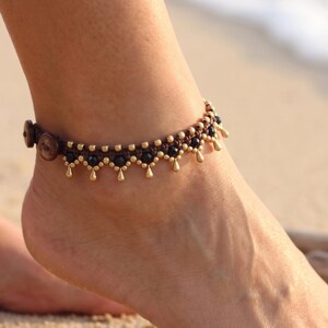 May include: An anklet featuring a dark brown cord base with small black beads and metallic gold-toned teardrop-shaped charms. The design includes a repeating pattern of beads and dangling gold accents, secured with a dark brown circular button closure.