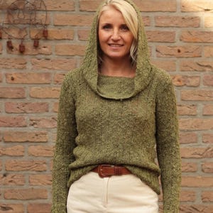 Könnte beinhalten: Eine Frau trägt einen grünen Kapuzenpullover mit einem Rollkragen und einem hellbraunen Gürtel. Sie trägt auch einen cremefarbenen Rock mit Schlitz. Der Hintergrund ist eine Backsteinmauer.