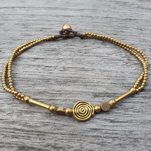 May include: A double-strand bracelet featuring small metallic gold-toned beads, two elongated cylindrical beads, two flat circular discs, and a central spiral-shaped charm. The bracelet is secured with a dark brown cord and a small spherical bead clasp.
