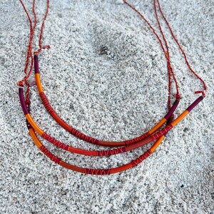 以下が含まれることがあります： 赤、オレンジ、黄色の3本の編組コード。コードの中央には濃い赤色の糸が織り込まれている。コードは白い砂浜の上に置かれている。