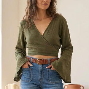 May include: An olive green long-sleeved wrap top with a V-neckline and flared sleeves paired with high-waisted blue denim jeans featuring front patch pockets and a brown woven leather belt with a gold-toned buckle at the waist.