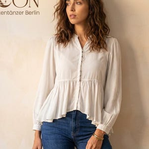 May include: A woman wearing a white long-sleeve blouse with a button-down front and a flared hemline, paired with blue jeans. The blouse has a subtle peplum detail. The image includes the text "FRIEDA MOON By Weltentänzer Berlin".