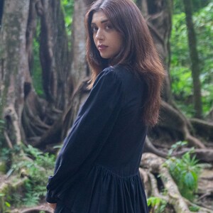 May include: A woman with long dark brown hair is wearing a black long-sleeved blouse with a ruffled hem. She is standing in a forest setting with large trees and roots.