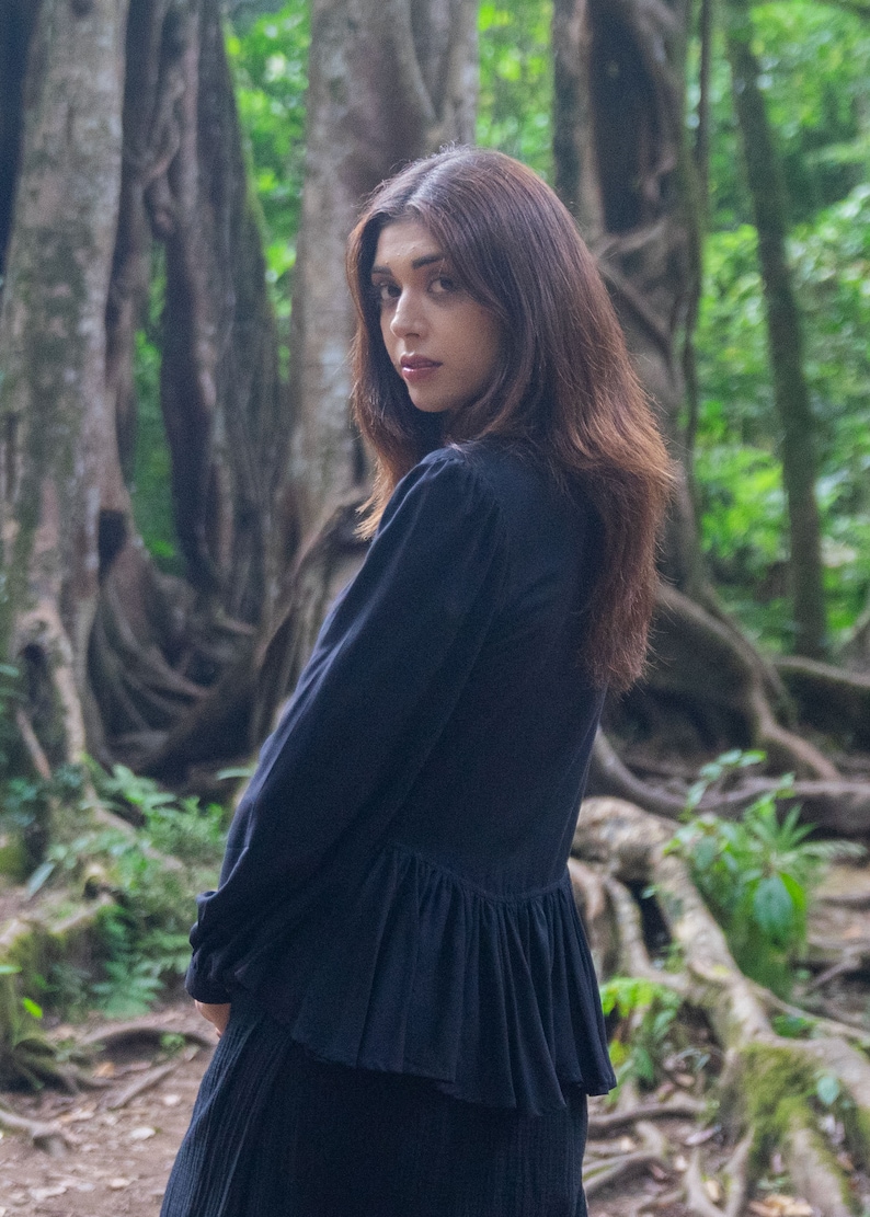 May include: A woman with long dark brown hair is wearing a black long-sleeved blouse with a ruffled hem. She is standing in a forest setting with large trees and roots.