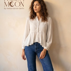以下が含まれることがあります： 白いボタンダウンのブラウスとフレア裾の女性、青いジーンズを着用。 ブラウスは長袖で、微妙な質感があります。 画像には「FRIEDA MOON By Weltentänzer Berlin」というテキストが含まれています。